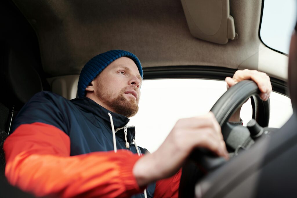 Low angle shot of a bearded man driving a car, wearing a hoodie and knit cap.